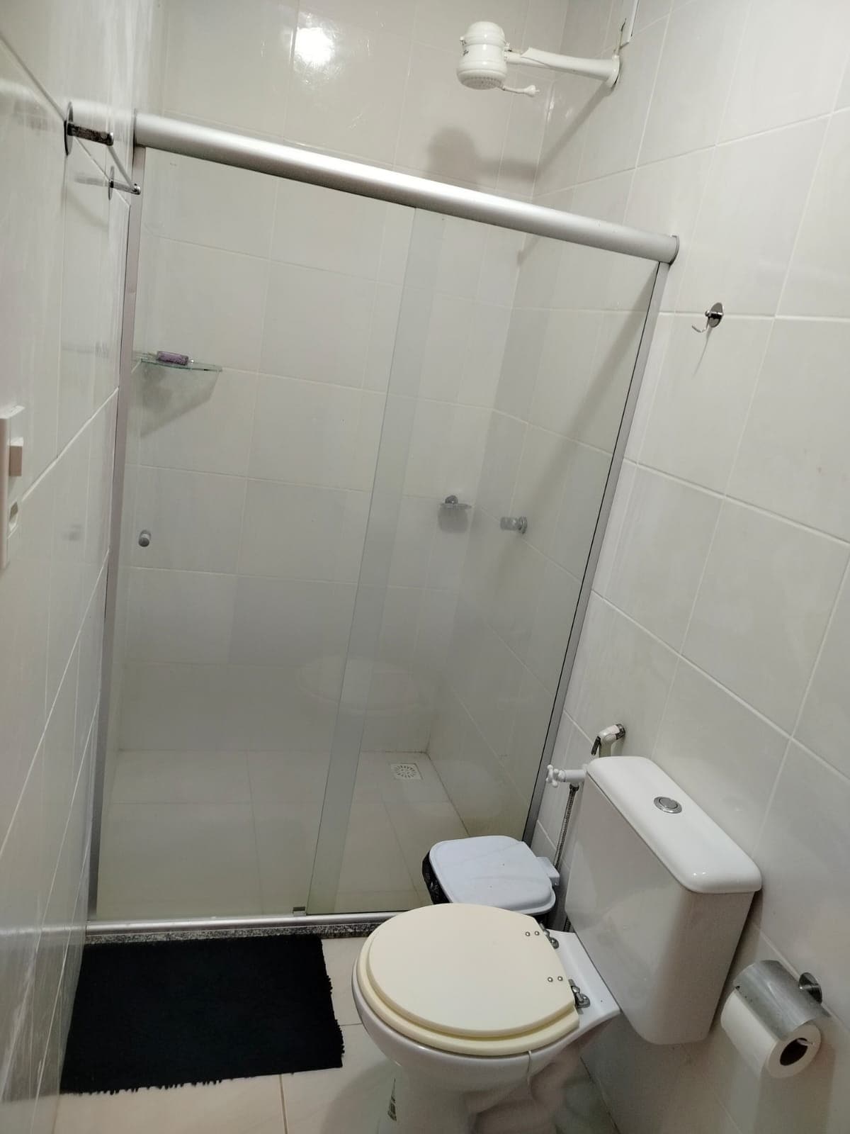The bathroom features a glass shower enclosure and a modern toilet. Clean white tiles line the walls, contributing to a bright and airy atmosphere. A simple black mat is positioned on the floor near the shower area, enhancing the space's practicality.
