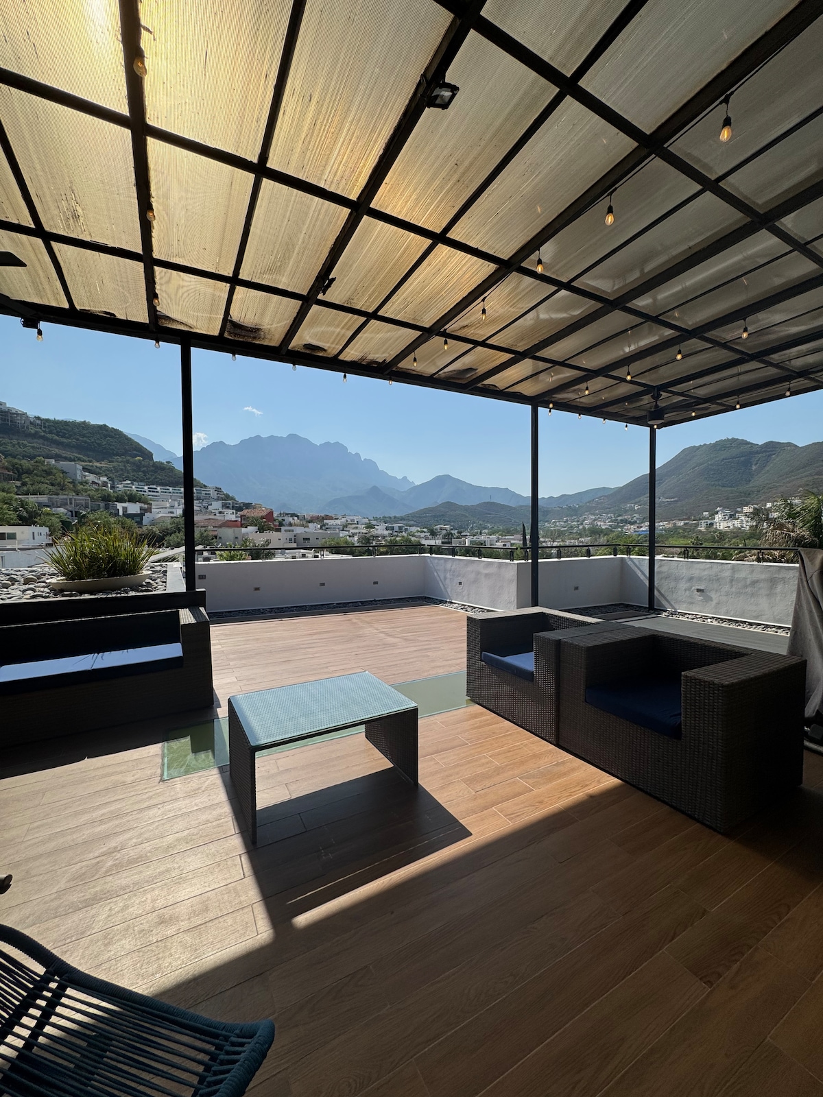 A spacious rooftop terrace features a covered seating area with a dark wicker seating arrangement and a low coffee table. The area is surrounded by large windows, providing a panoramic view of the mountains and nearby buildings. Soft lighting enhances the tranquil atmosphere.