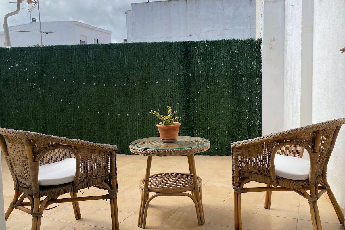 A private terrace features a small glass table surrounded by two woven chairs with soft cushions. A potted plant sits at the center of the table, complemented by a lush green backdrop, providing a serene outdoor space for relaxation.