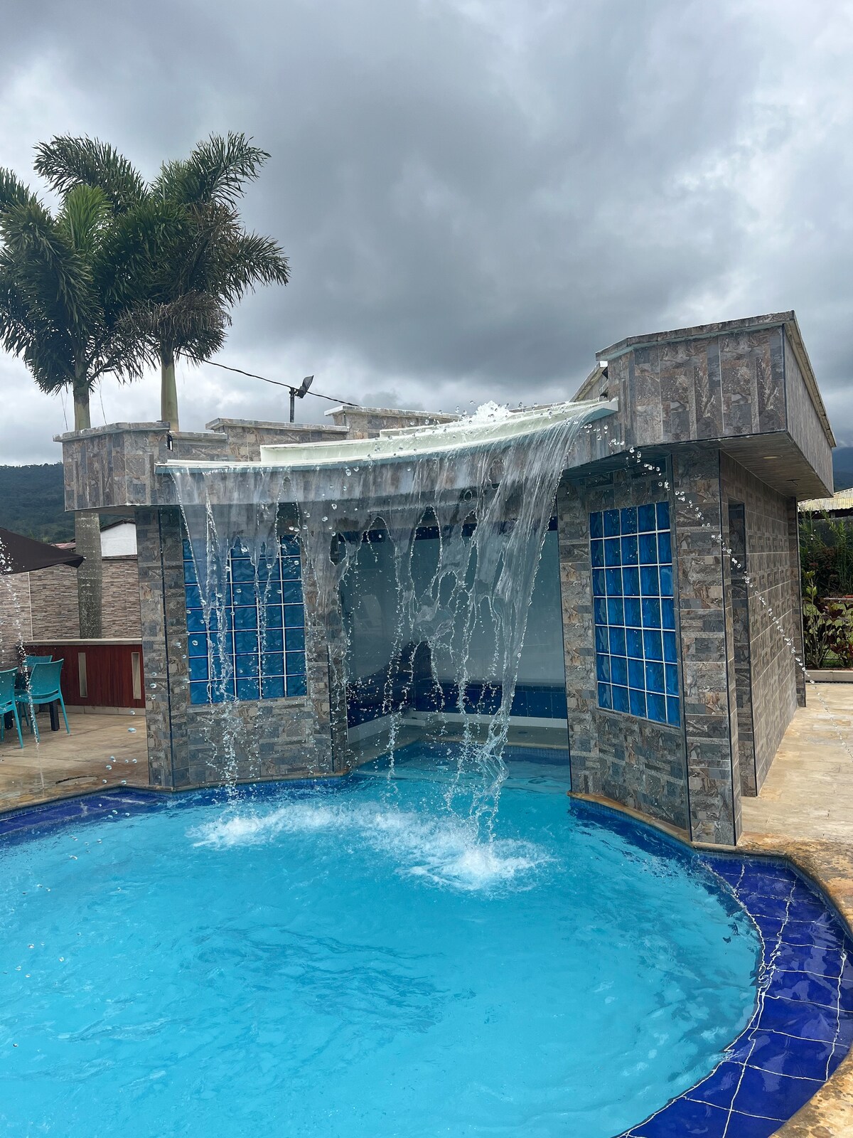 A clear blue pool features a waterfall cascading from a modern stone structure. The pool's edge is outlined with decorative tiles, and tropical palm trees are visible in the background, contributing to a serene environment.
