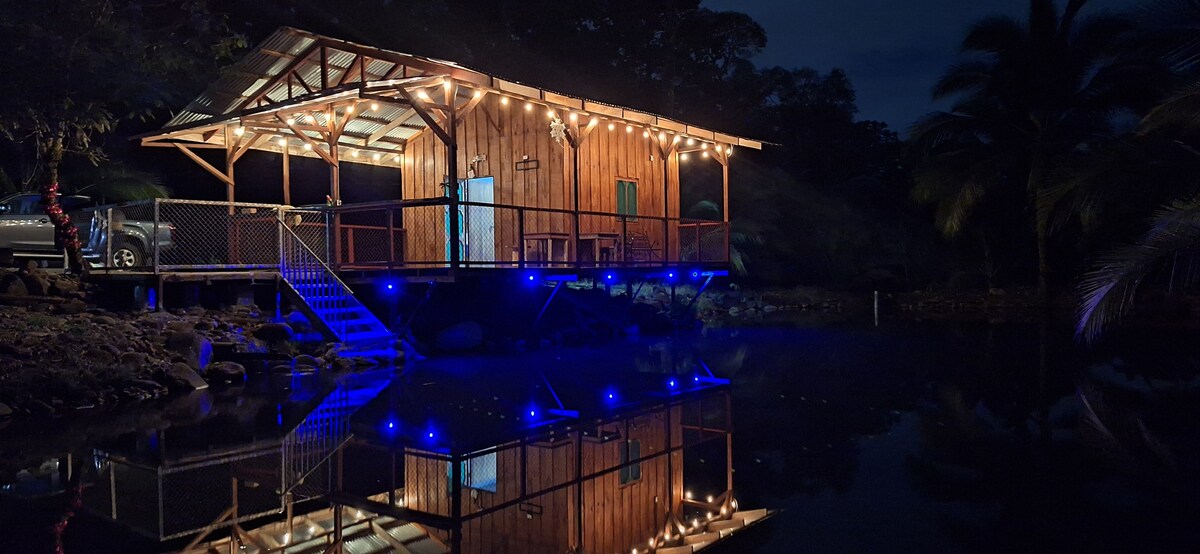 A wooden cabin is set against a tranquil pond, illuminated by soft string lights along the eaves. The water reflects the structure and lights, creating a serene atmosphere. Surrounding palm trees can be seen in the evening backdrop, enhancing the calm setting.