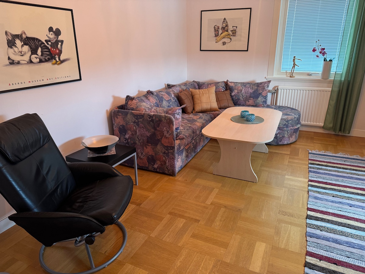 A cozy living area is furnished with a floral patterned sofa and a light wooden coffee table. A comfortable black armchair is positioned nearby. Two framed artworks adorn the walls, and natural light filters through a window with light curtains, highlighting the wooden floor.