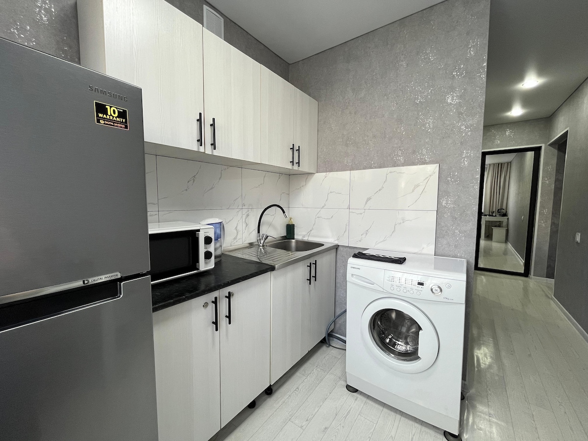A compact kitchen is fitted with modern cabinetry in light tones. A stainless steel refrigerator and a microwave are visible, alongside a sink and a washing machine. Sleek white countertops reflect natural light, contributing to a clean and functional workspace.