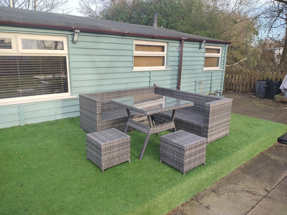 A garden seating area is presented featuring a glass-topped table surrounded by four cushioned chairs and additional stools. The area is bordered by well-maintained artificial grass, with a backdrop of light blue shiplap siding from the cabin.