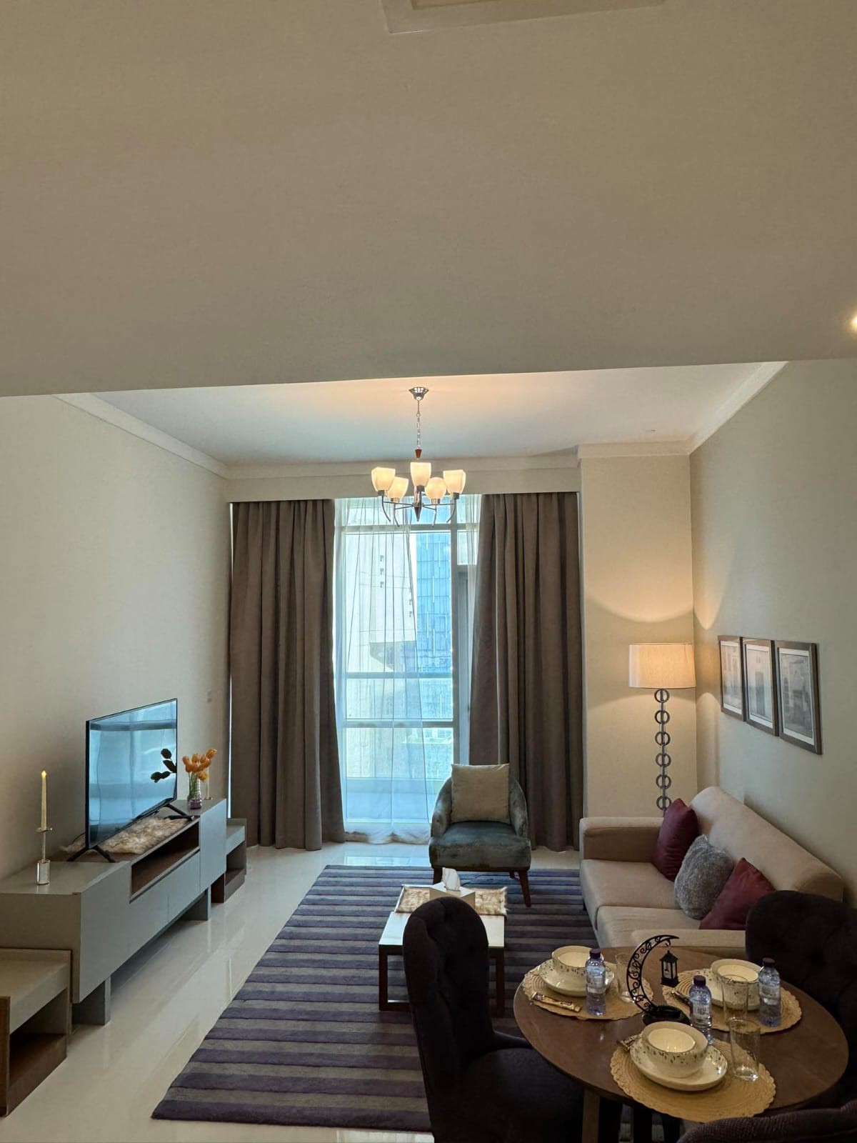 A well-lit living space is highlighted by a soft sofa and an accent chair, facing a modern television. A dining table is set for four, accompanied by elegant tableware. Floor-to-ceiling drapes frame a large window, allowing natural light to fill the room.