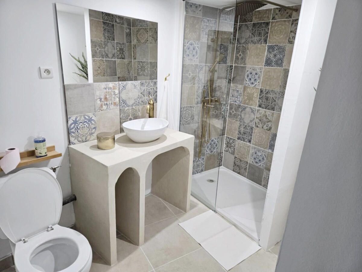 A modern bathroom is presented, featuring a minimalist sink atop a sculptural beige pedestal. Colorful patterned tiles adorn the shower wall, while a clear glass enclosure offers an open feel. The toilet is positioned nearby, and a neutral tile floor completes the space.