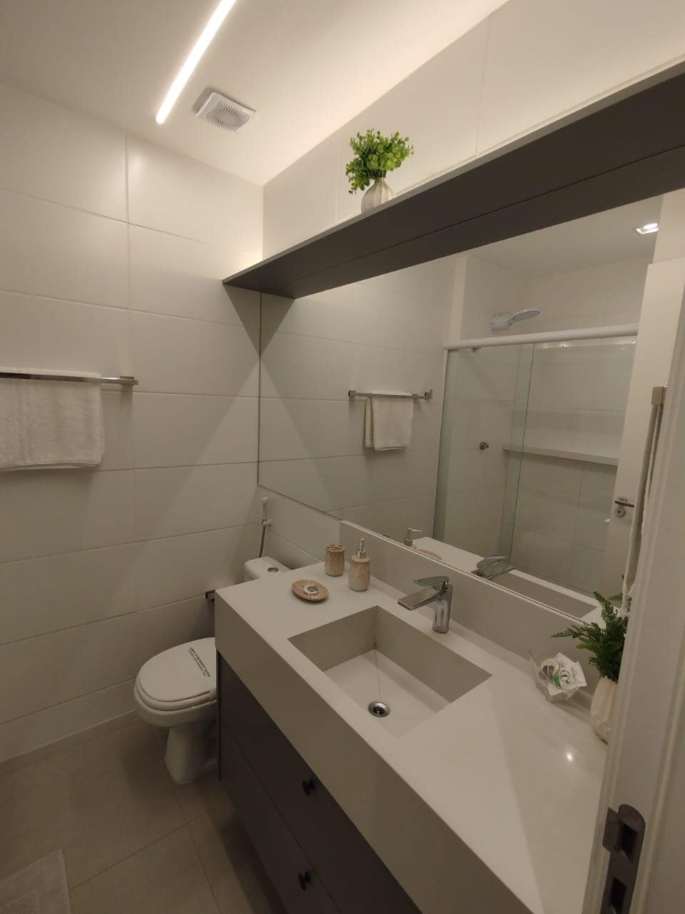 A modern bathroom features a sleek sink with a minimalist countertop and a large mirror above. Soft monochromatic tiles adorn the walls. A glass shower enclosure is visible, alongside essential toiletries arranged on the counter. Towels are neatly hung on wall-mounted racks.