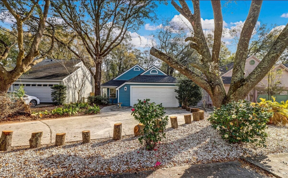 The exterior of a single-family home is visible, featuring a light blue façade and a well-maintained driveway. Large trees frame the front yard, and trimmed bushes with pink flowers add color to the landscape. The overall setting appears serene and welcoming.