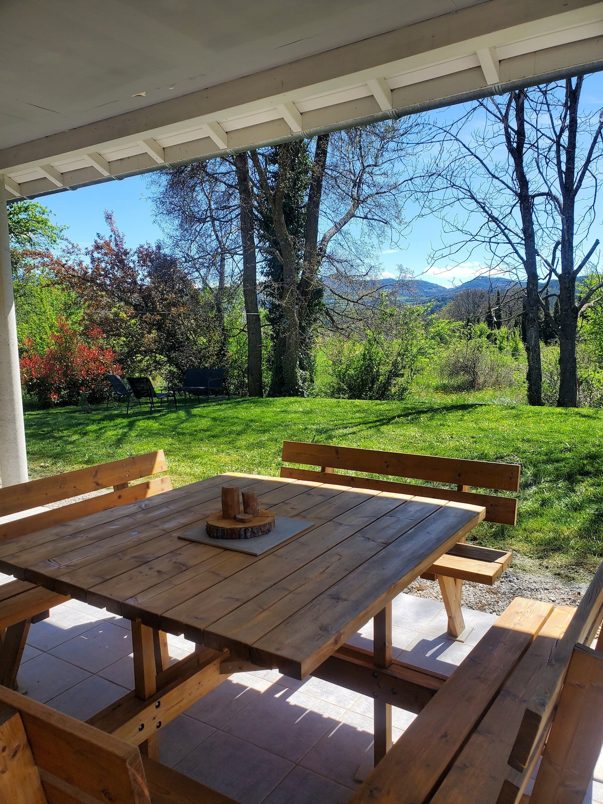 A wooden dining set is positioned on a patio, featuring a square table surrounded by benches. Lush green grass and trees can be seen in the background, with hints of distant mountains poking through the foliage under a clear blue sky.