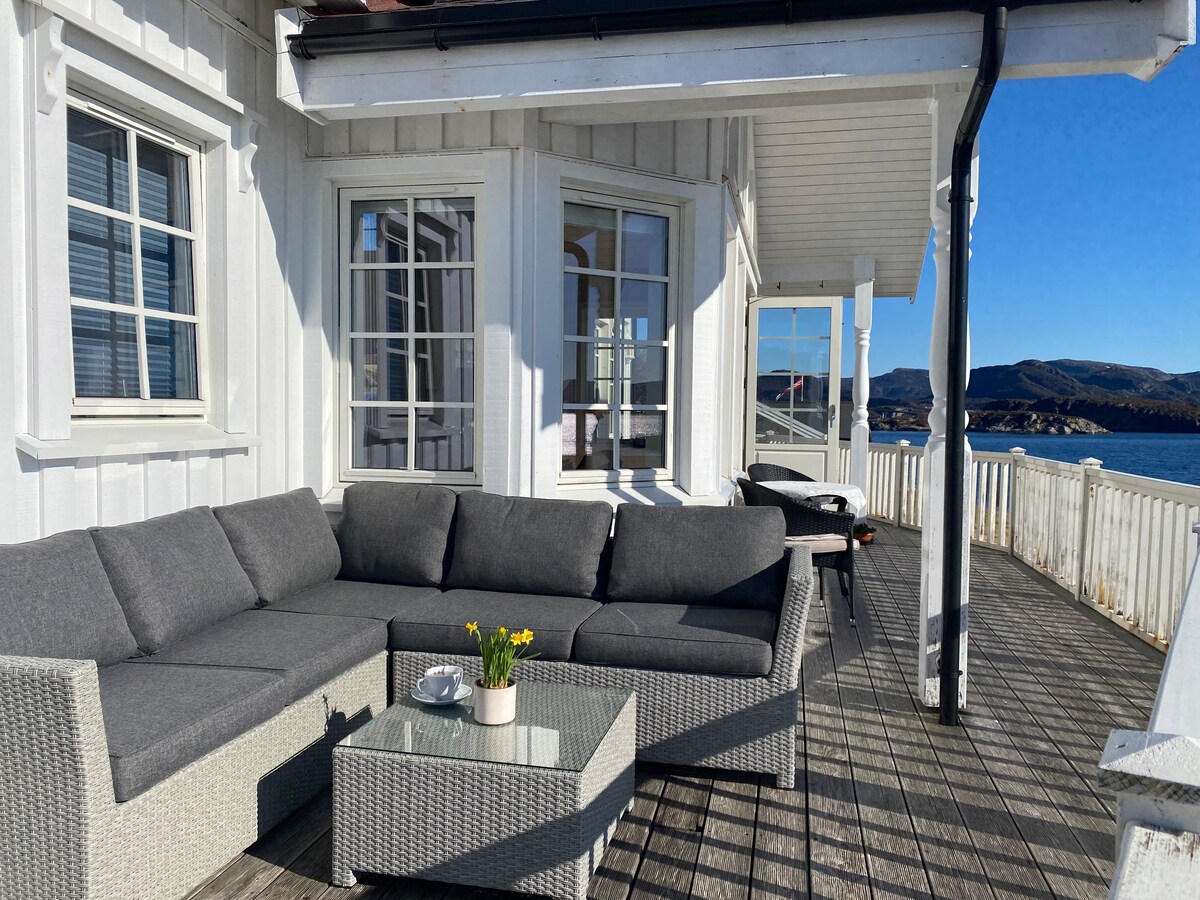 A spacious outdoor seating area features a gray sectional sofa and a glass-top table with a small potted plant. Large windows provide ample natural light, and a scenic view of the water and surrounding nature is visible in the background.