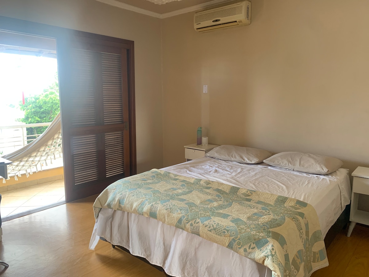A spacious bedroom is featured, showcasing a large bed with light bedding. A bedside table is positioned on each side. Natural light enters through a sliding door leading to a balcony, where a hammock is visible, enhancing the room's inviting atmosphere.