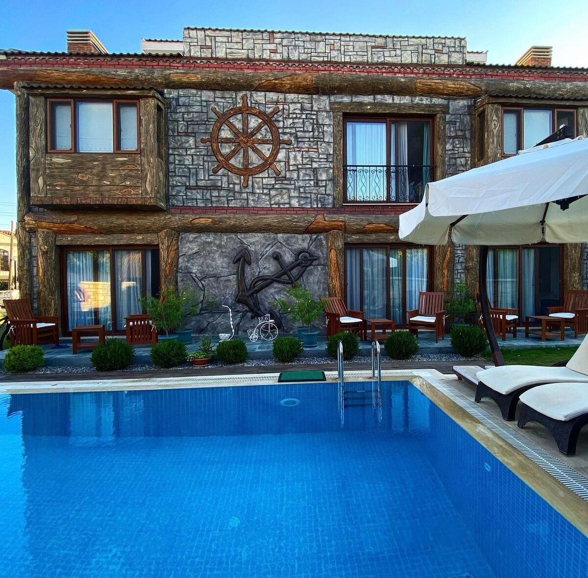 The exterior of a two-story stone building is showcased, featuring large windows and a nautical theme with a ship's wheel decoration. In the foreground, a clear blue pool is visible, alongside lounging chairs arranged on a wooden deck, surrounded by greenery.