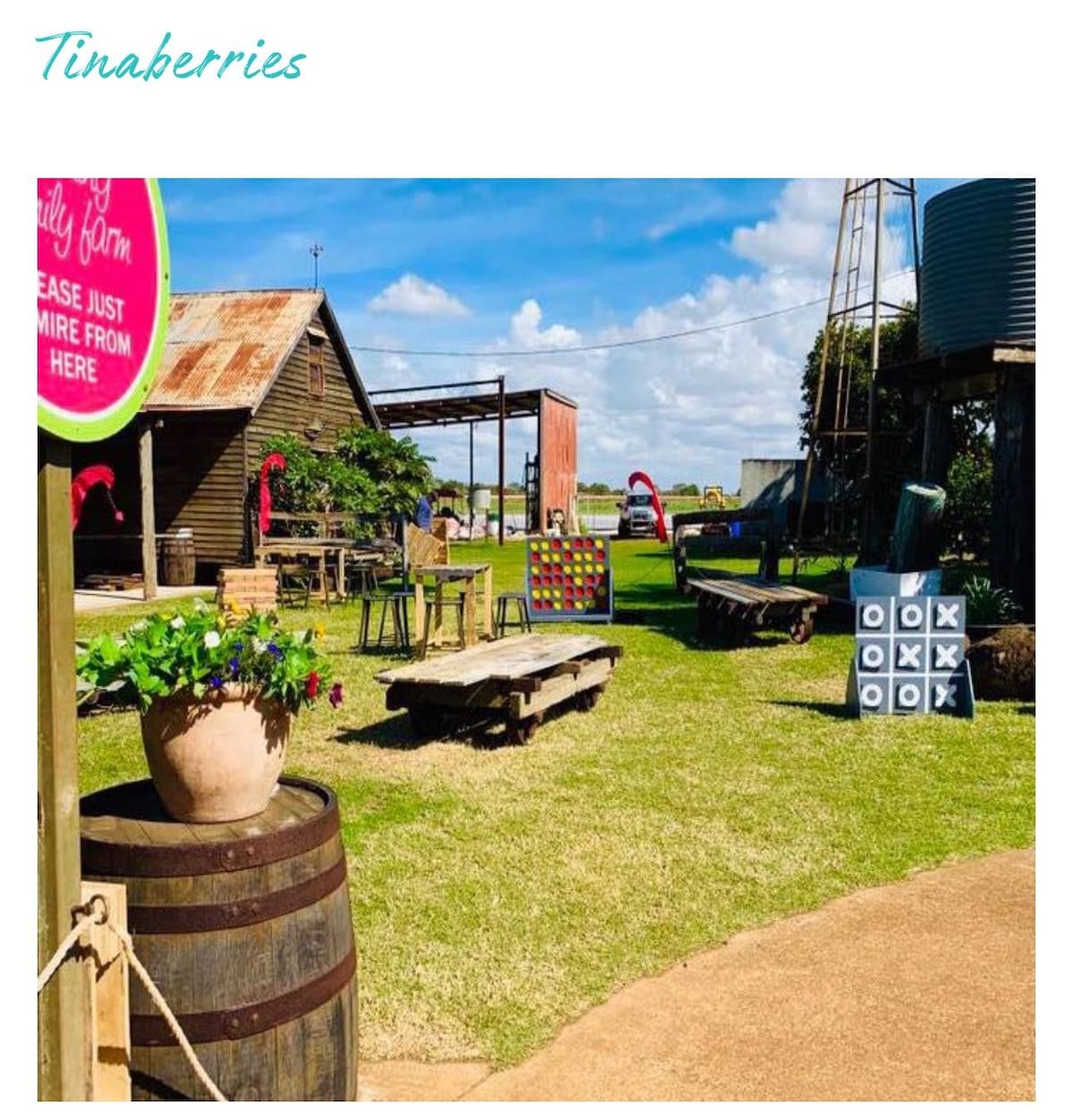 An outdoor space is depicted with wooden benches and picnic tables set on green grass. Colorful games are placed strategically, and a rustic building is visible in the background. Clear blue skies enhance the open atmosphere of the inviting setting.
