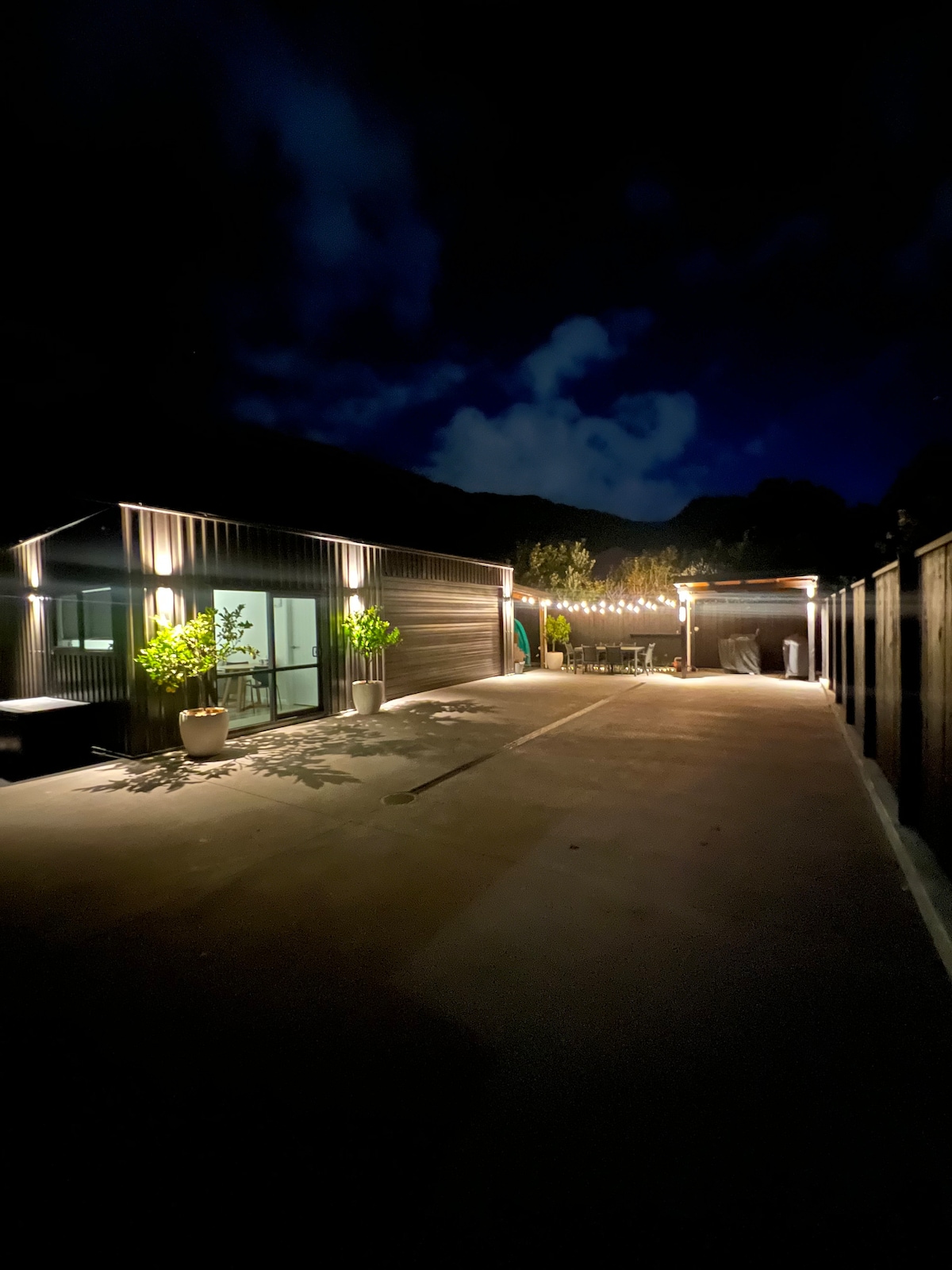 A private driveway is illuminated by warm lights, leading to a stylish building with glass windows. Potted plants add a touch of greenery, while a gathering area with outdoor seating is visible in the distance. The serene night sky is adorned with clouds.