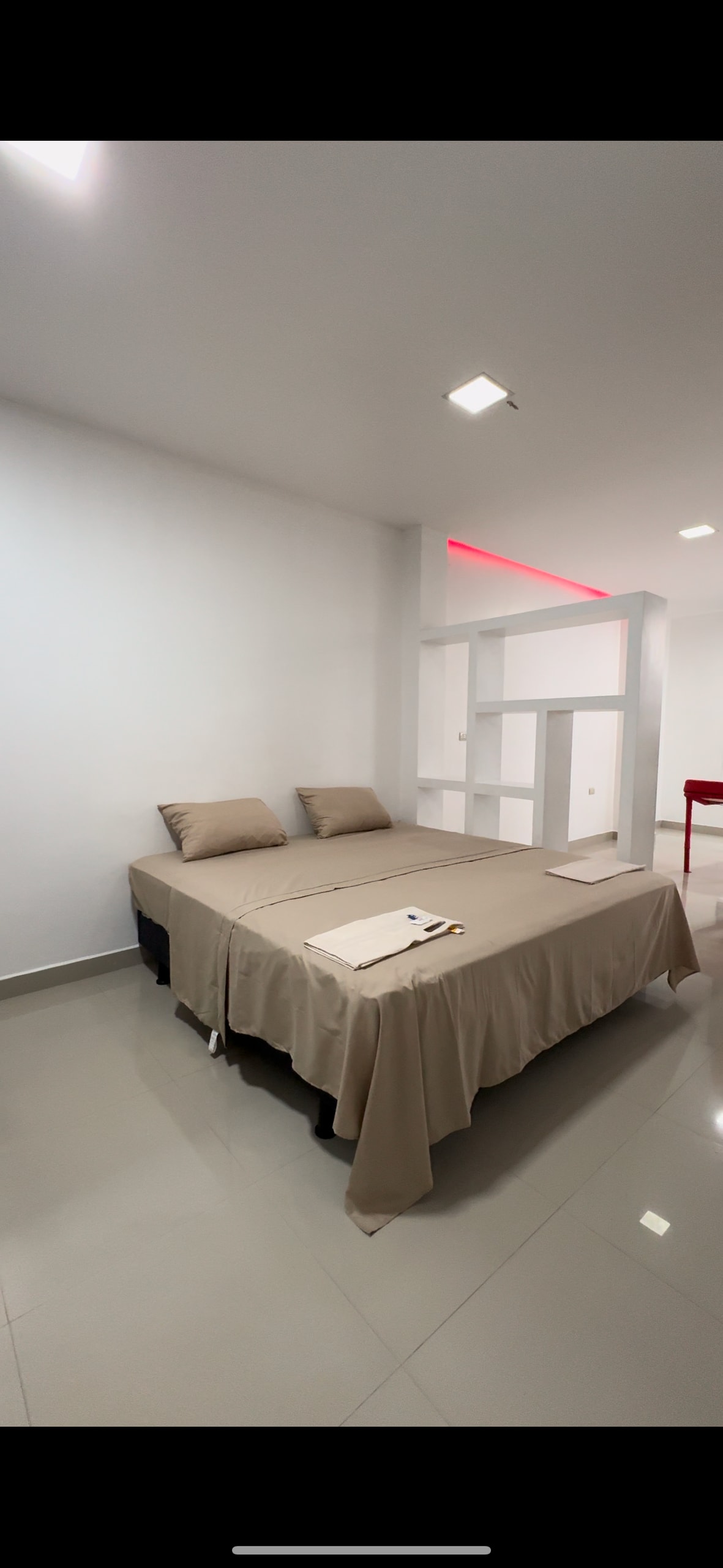 A spacious bed is dressed in neutral-toned linens, accompanied by two matching pillows. The room features smooth, reflective tile flooring and a minimalistic design, with soft ceiling lighting providing overall illumination. A simple wall unit is visible in the background, enhancing the room's uncluttered aesthetic.