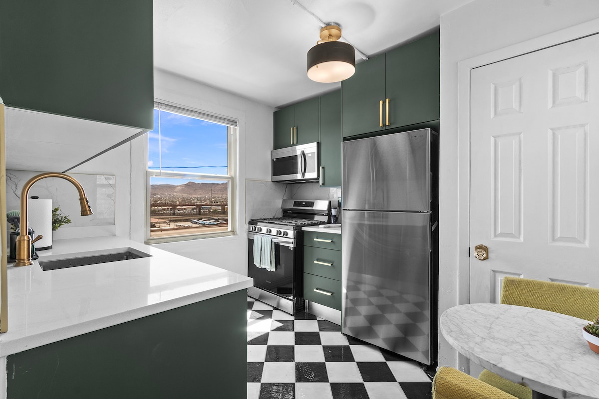 A modern kitchen is showcased with stylish green cabinets and sleek stainless steel appliances. The black and white checkered floor adds a classic touch. Natural light floods in from a nearby window, illuminating the counter and dining area with a round marble table and two yellow chairs.