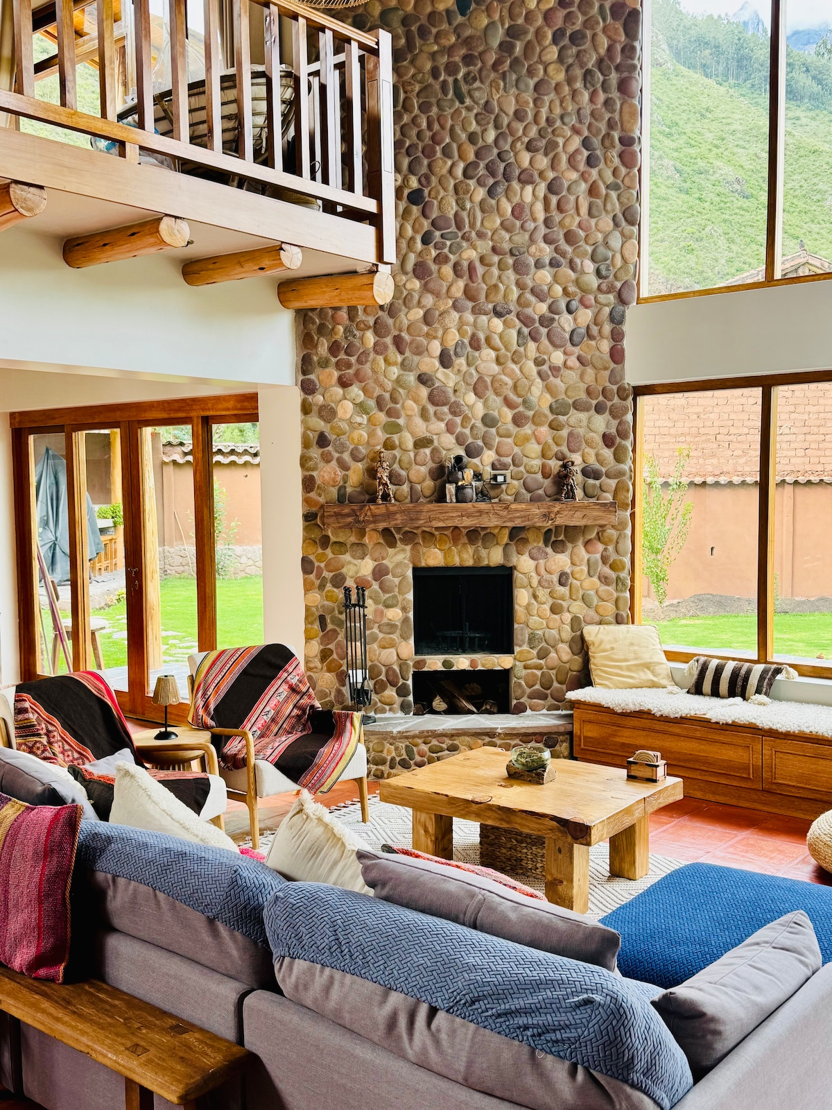 A spacious living area features a stone fireplace as a focal point against a backdrop of large windows. Comfortable seating options include couches adorned with varied textiles and several armchairs. Natural light fills the space, highlighting the warm tones of the decor and surrounding greenery.