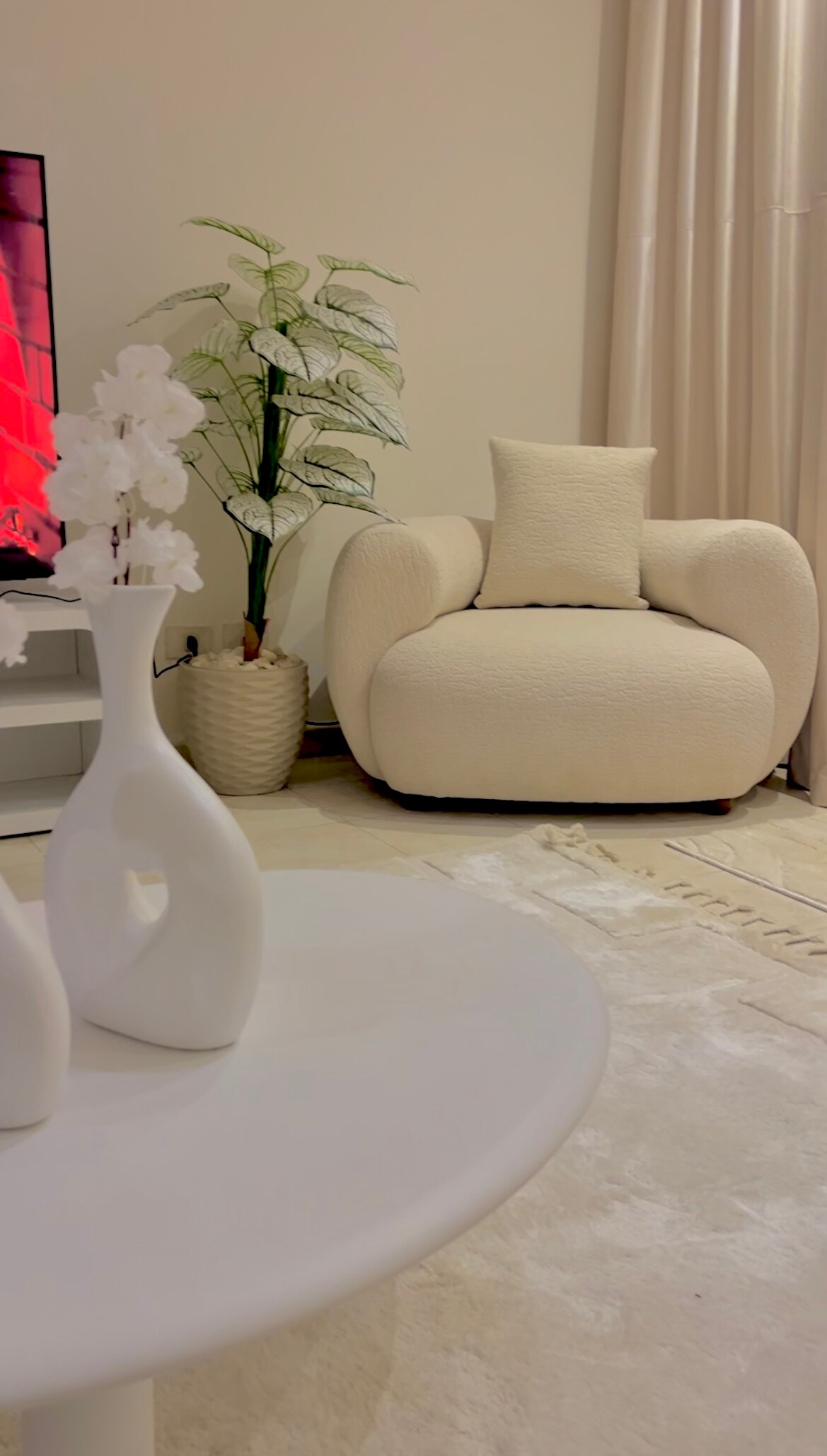 A soft-textured armchair in a light color is positioned beside a tall potted plant. A white vase containing delicate flowers sits on a round coffee table, which is placed on a plush area rug, complementing the overall tranquil decor.