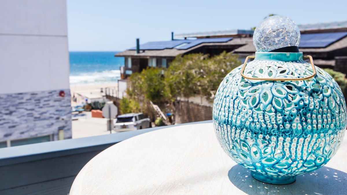 A decorative blue glass lantern rests on a light-colored table, with the ocean in the background. The vibrant blue of the lantern contrasts with the natural hues of the surrounding landscape and the sandy beach extending towards the water.