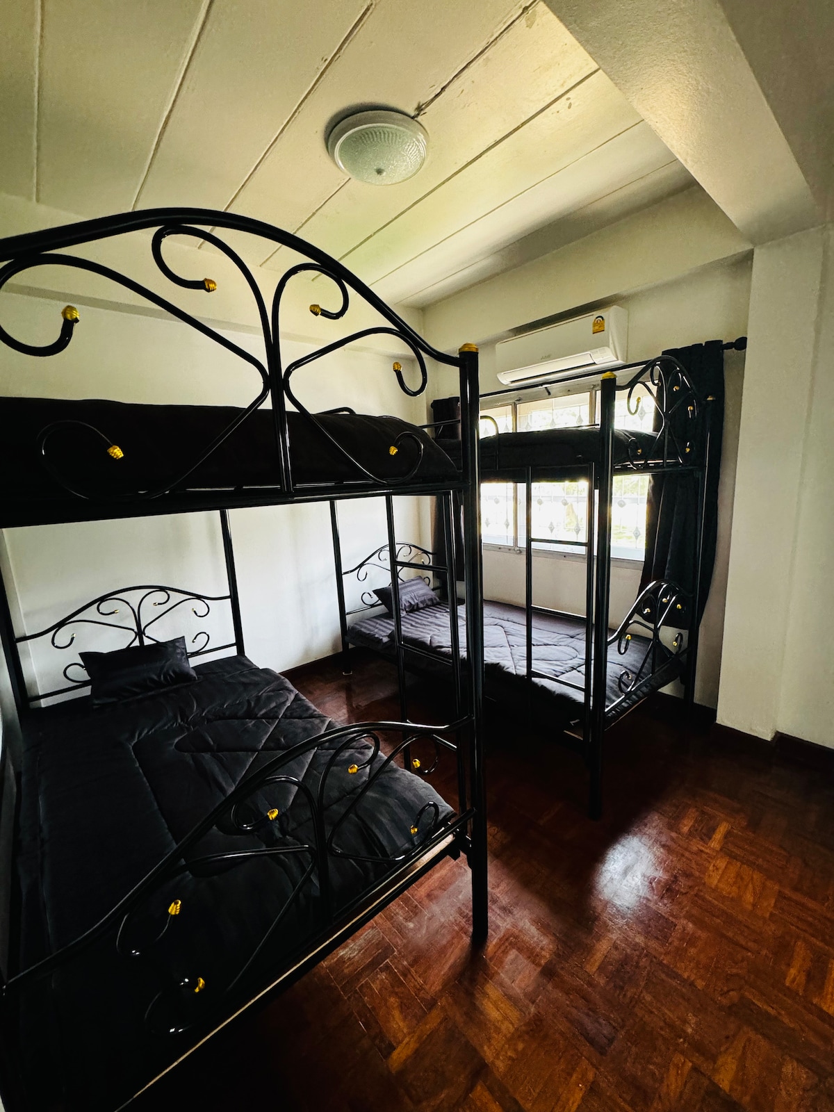 A shared sleeping area features two sets of bunk beds with black metal frames and dark bedding. Natural light enters through a window, and the wooden floor enhances the room's overall warmth. A ceiling fan is positioned above, providing ventilation.