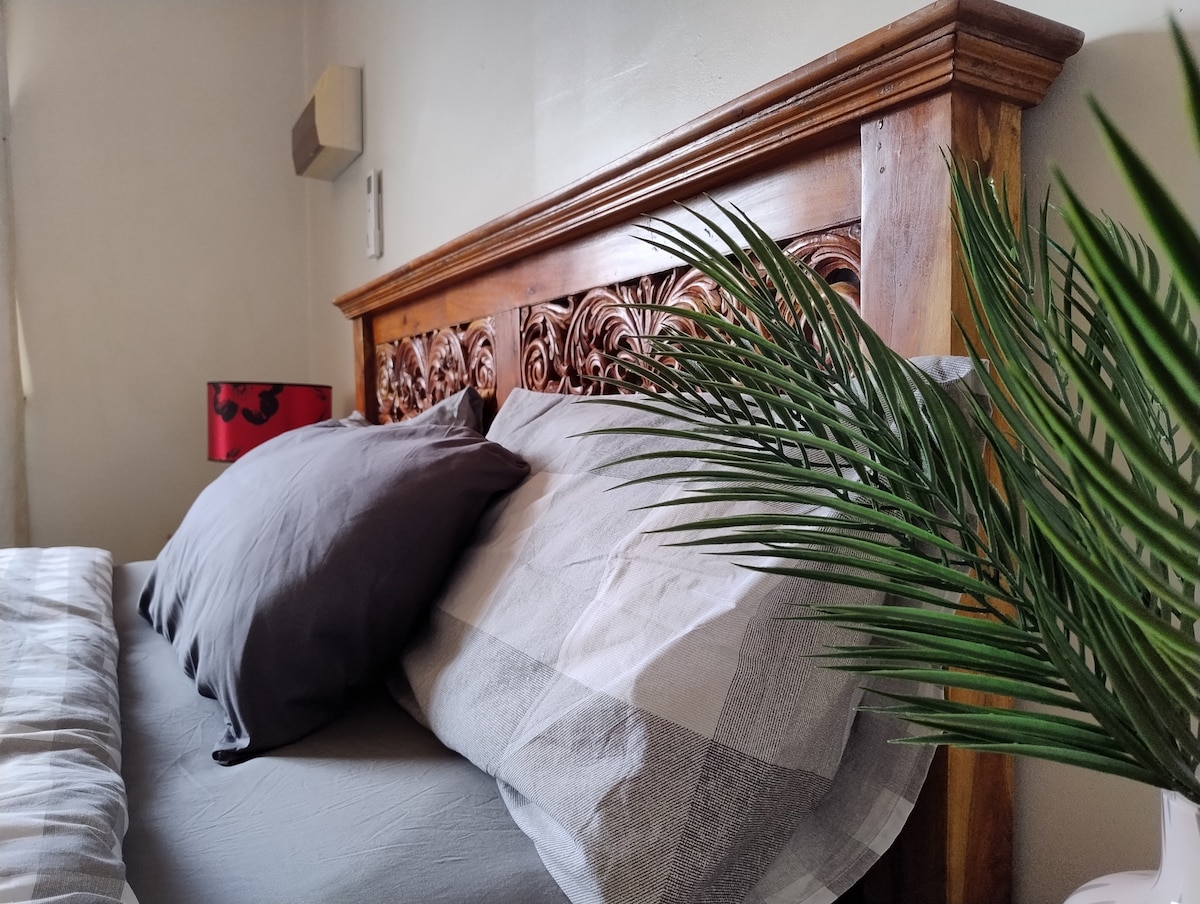A close-up view of a spacious bed featuring a carved wooden headboard. Two pillows are arranged on top of a soft, checkered blanket. A small decorative lamp with a red shade is visible beside the bed. A green plant adds a natural touch to the scene.