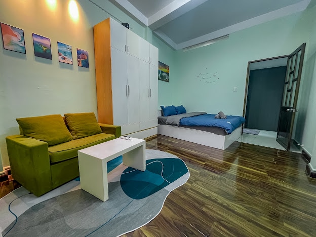A well-lit room features a cozy seating area with a green couch facing a small white table. A spacious bed with blue linens is positioned against the wall, adjacent to a closet with sliding doors. Artistic photographs are displayed above the couch.