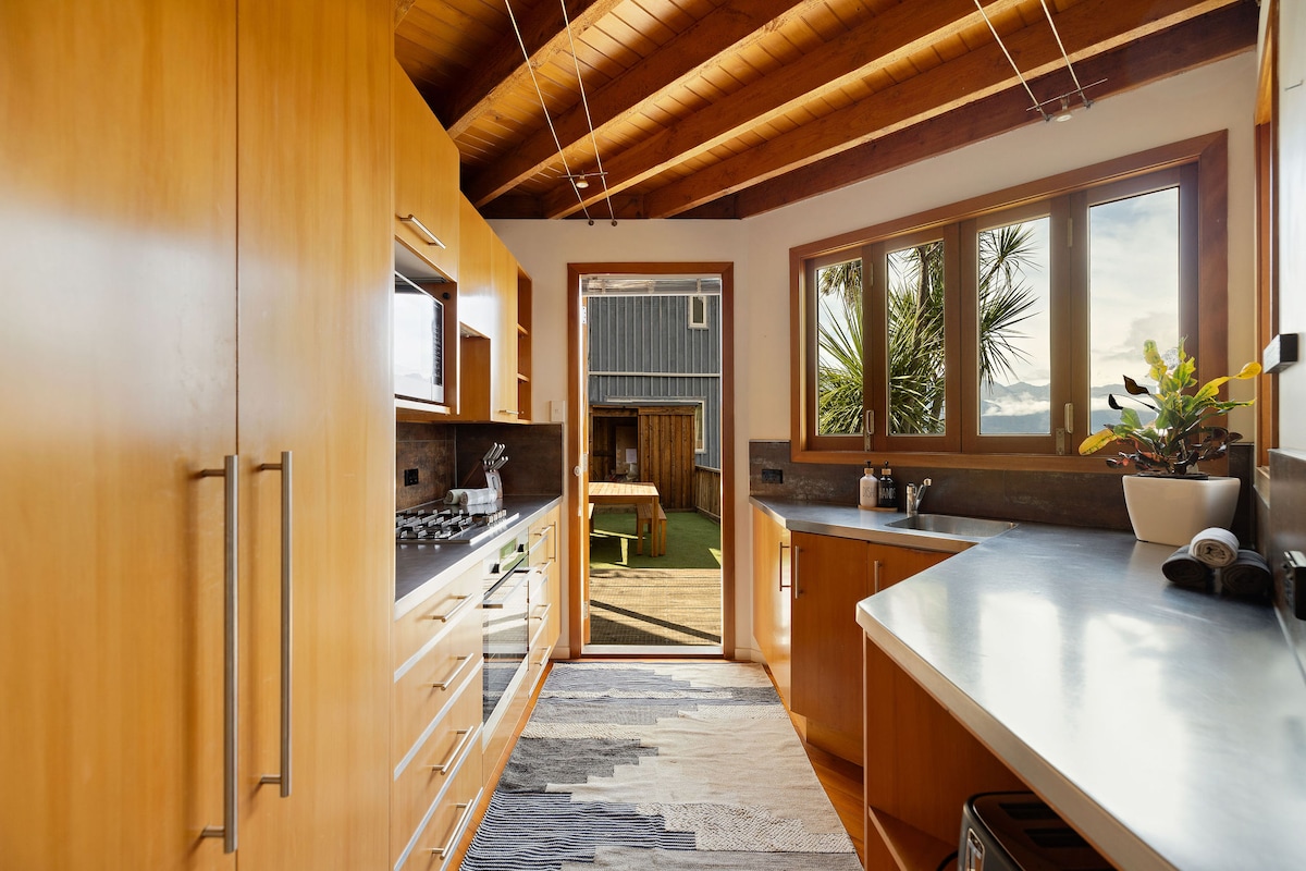 The kitchen features ample cabinetry with warm wood tones and modern appliances. Large windows allow natural light to fill the space, while a door leads to an outdoor area. A textured rug is placed on the floor, adding warmth to the design.
