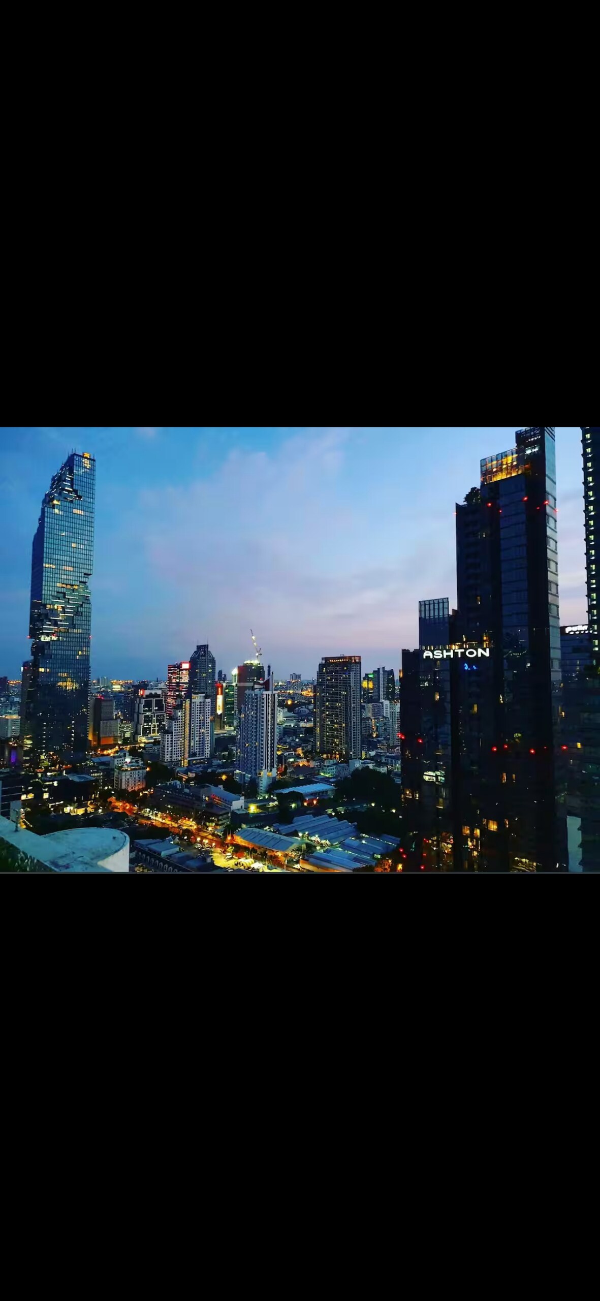 A city skyline is captured at dusk, showcasing a mix of modern skyscrapers and vibrant street lights. The building heights vary significantly, with a prominent tower illuminated by lights against the evening sky.