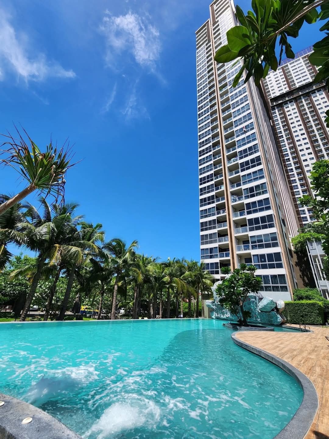 A wide view showcases the inviting swimming pool, surrounded by lush greenery and palm trees. The 45-story building towers in the background, reflecting the clear blue sky. Water splashes gently create a serene sound against the smooth pool surface, enhancing the tranquil outdoor space.