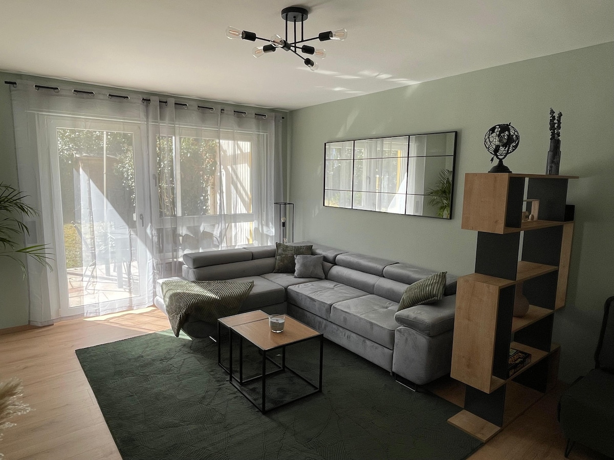 A modern living area is presented with a gray sectional sofa and a glass coffee table. Soft natural light enters through large sliding doors that lead to an outdoor terrace. A minimalist bookshelf is positioned against the green wall, complementing the overall calm color palette.