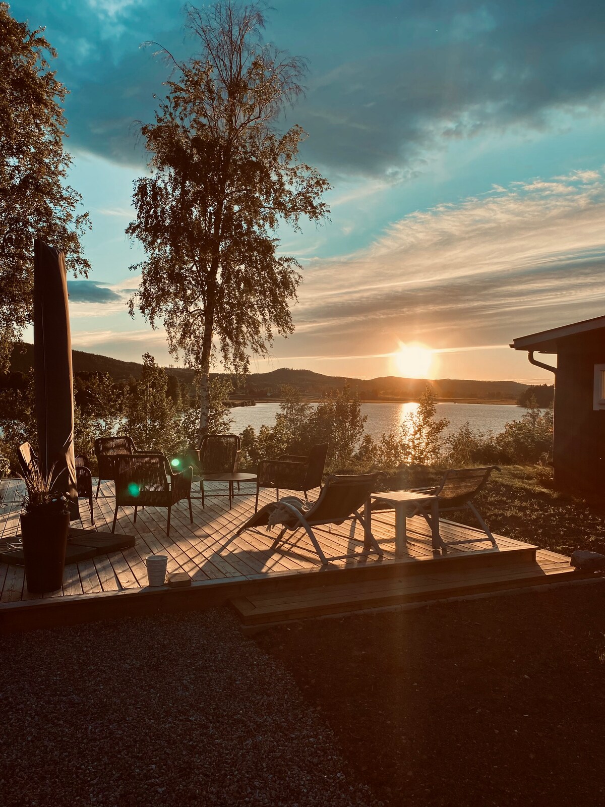 A deck is positioned with several chairs and a small table, offering a view of the water and surrounding landscape. A tall umbrella provides shade. The warm glow of the sunset casts a gentle light across the scene, reflecting off the water's surface.