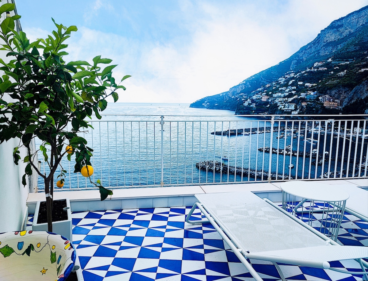 A terrace features stunning sea views with geometric patterned flooring in blue and white. A lemon tree adds a touch of greenery. Two sun loungers are positioned overlooking the water, inviting relaxation in a serene setting.