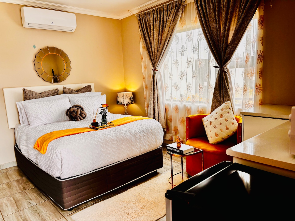 A well-appointed bedroom features a spacious bed dressed in neutral linens with an accent throw. Bright curtains frame a large window, allowing natural light to fill the room. A cozy seating area is provided with a stylish chair, and decorative elements enhance the tranquil setting.