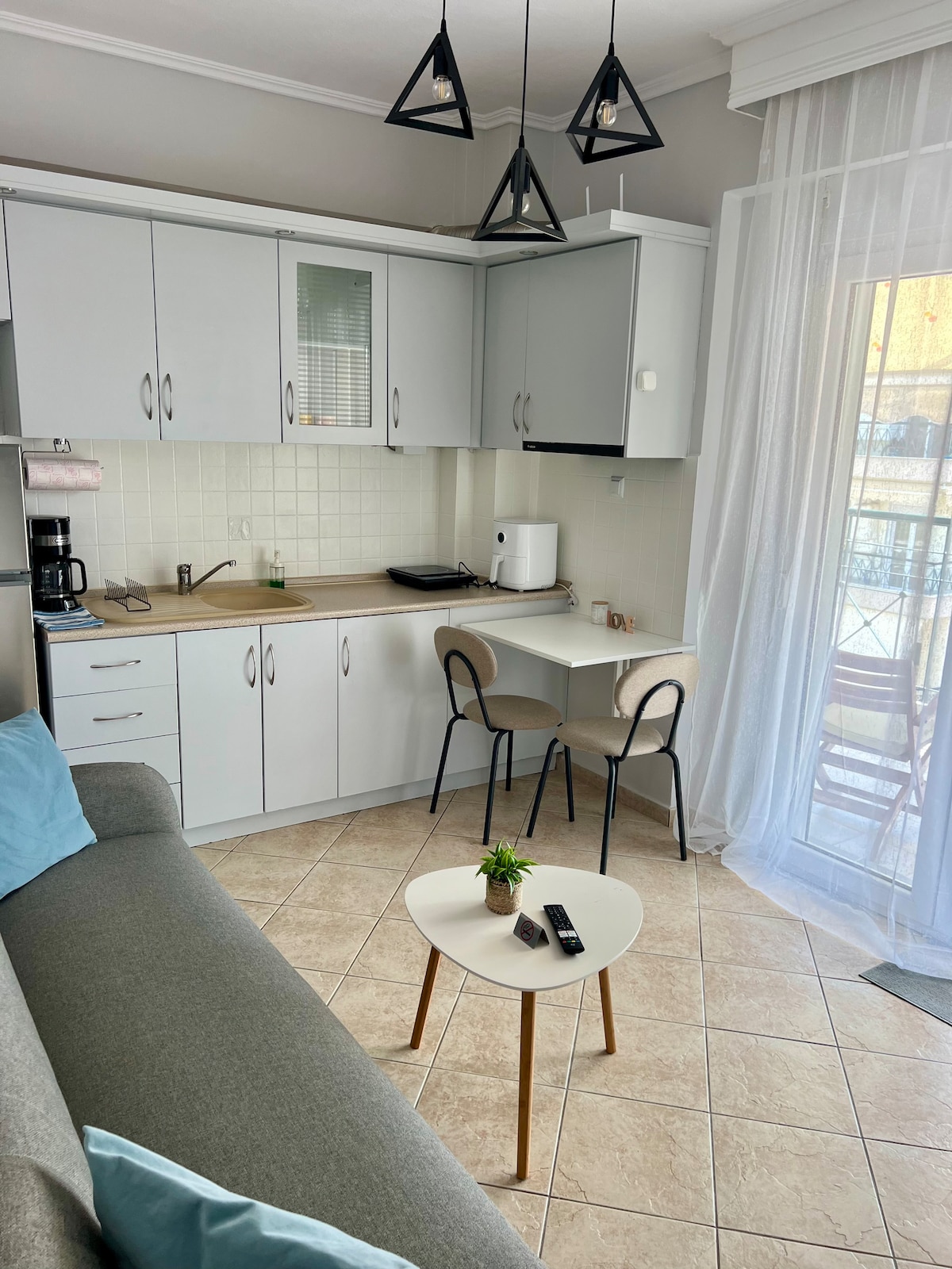 A bright living space features a compact kitchen with light gray cabinetry and modern appliances. A small dining area with two chairs is set beside a round table. Natural light enters through a sheer curtain, illuminating a comfortable sofa upholstered in soft gray.