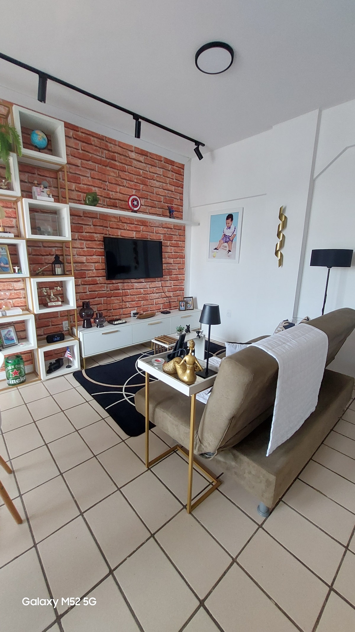 A modern living area features a sectional sofa against a brick accent wall, with a wall-mounted television above a low white media console. A small side table holds decorative items, and a floor lamp provides additional lighting. Geometric patterns in the rug complement the space.