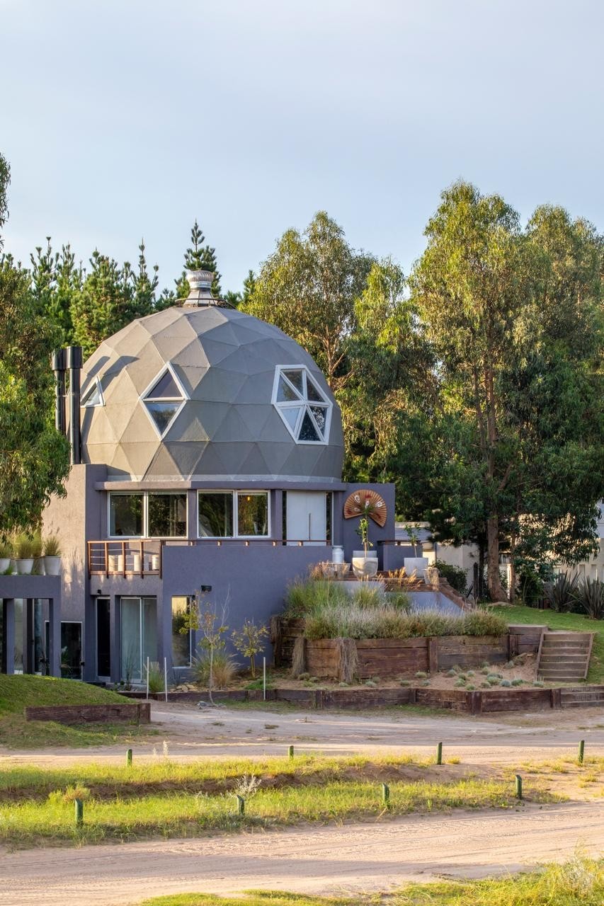 A unique domed structure is surrounded by lush greenery, showcasing multiple large windows. The building features a combination of natural and modern elements, with a wooden sculpture visible on the front terrace and a staircase leading to the entrance.