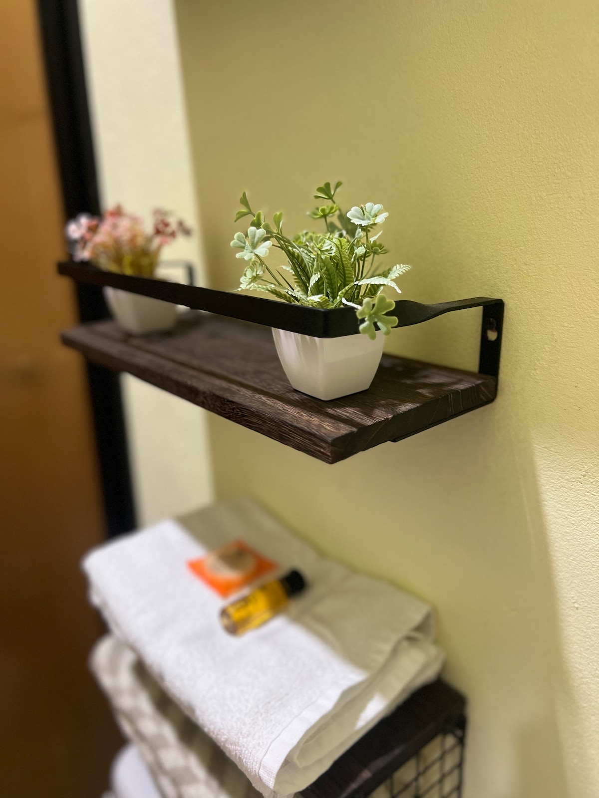 A wooden shelf is mounted on the wall, displaying two small potted plants. Below the shelf, neatly folded white towels are arranged, accompanied by a small bottle and a travel-size product resting on top.
