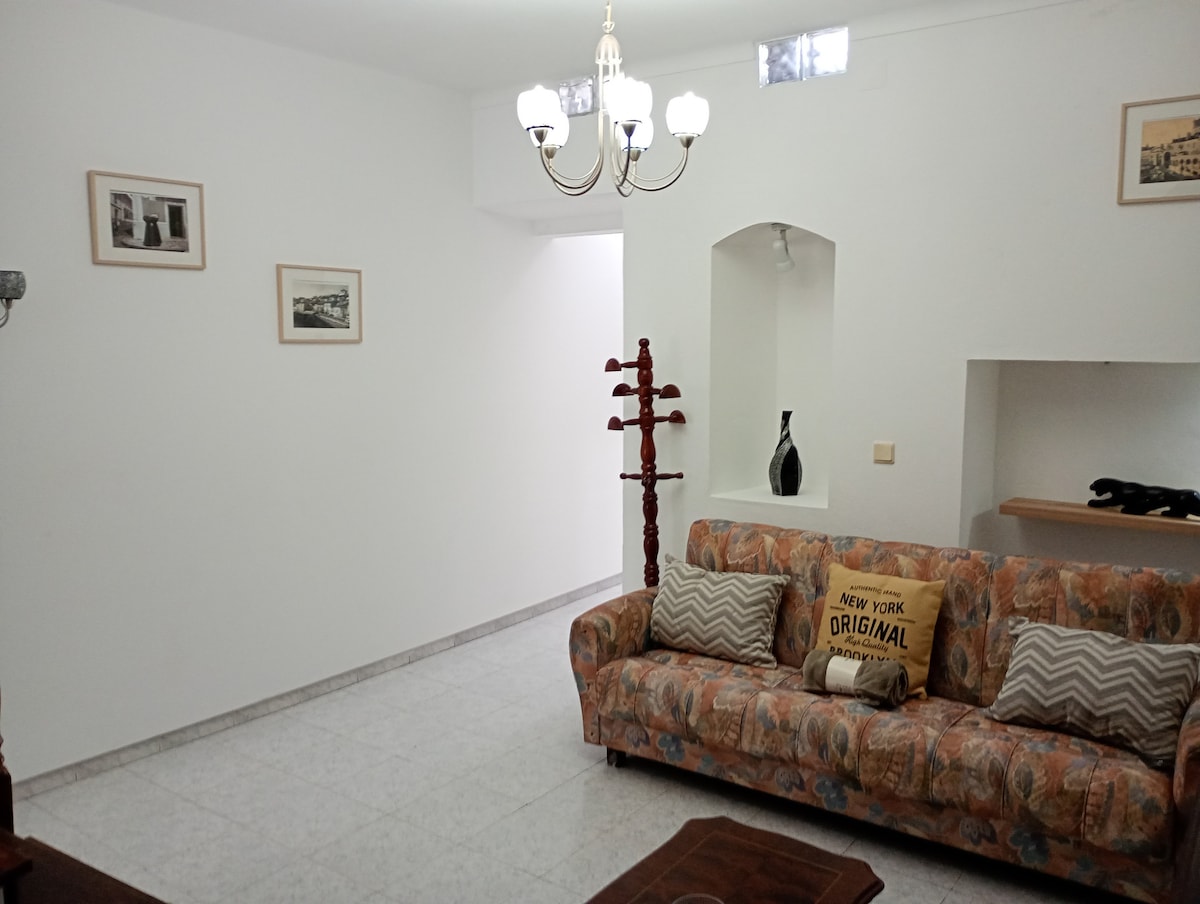 A bright living area is displayed, featuring a patterned sofa adorned with decorative cushions. A chandelier hangs from the ceiling, providing illumination. Framed artworks are mounted on the walls, while a small shelf and a decorative vase are positioned in the background.