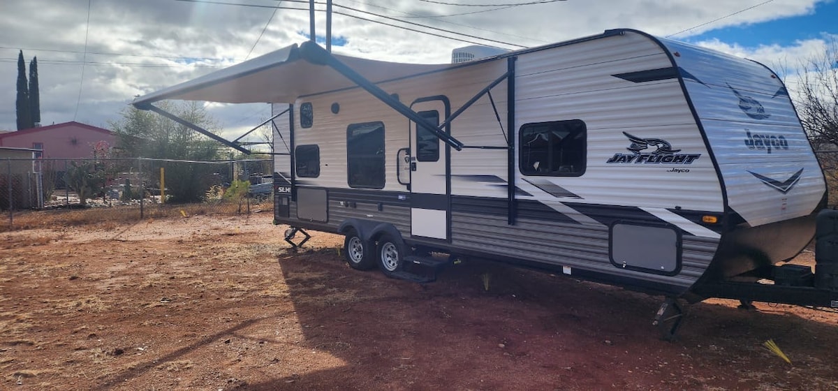 A travel trailer is prominently displayed in an outdoor setting, featuring a retractable awning extended over a spacious side. The exterior design presents a blend of dark and light grey colors, with a logo visible on the side, surrounded by a rugged landscape.