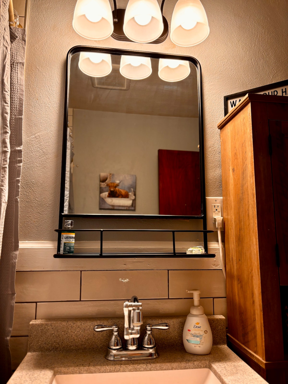 A bathroom sink area is visible, with a sleek countertop and a wall-mounted mirror featuring a black frame. The sink includes a modern faucet, while a bottle of hand soap sits nearby. Soft lighting is provided by three curved fixtures above the mirror.