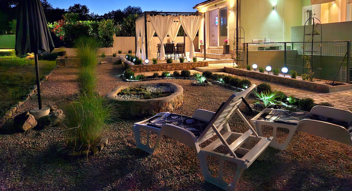 A landscaped outdoor area is illuminated in soft lighting, featuring a gravel pathway and various plants. Comfortable lounge chairs are positioned for relaxation. A dining area with a canopy is visible in the background, accompanied by a decorative stone feature in the garden.