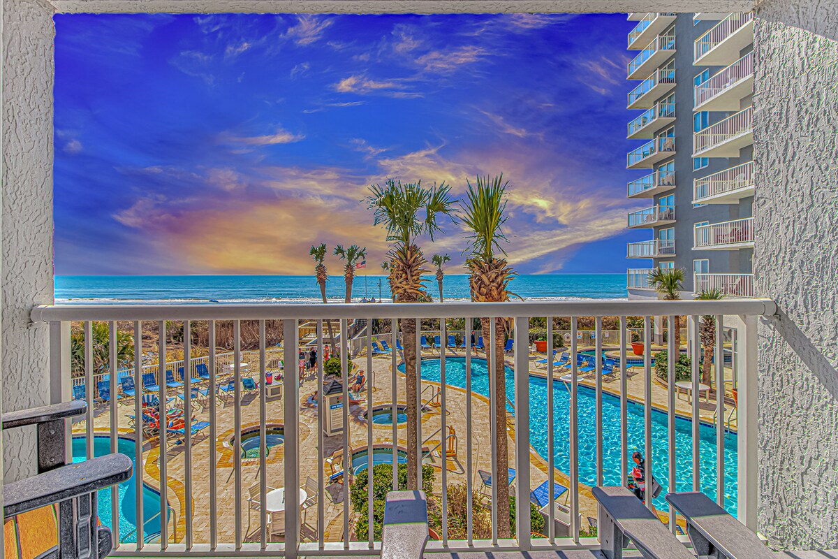 An oceanfront balcony offers a sweeping view of the Atlantic Ocean against a colorful sky. Below, a resort pool area is surrounded by palm trees and lounge chairs. Two comfortable chairs are positioned on the balcony, creating a perfect space for relaxation.