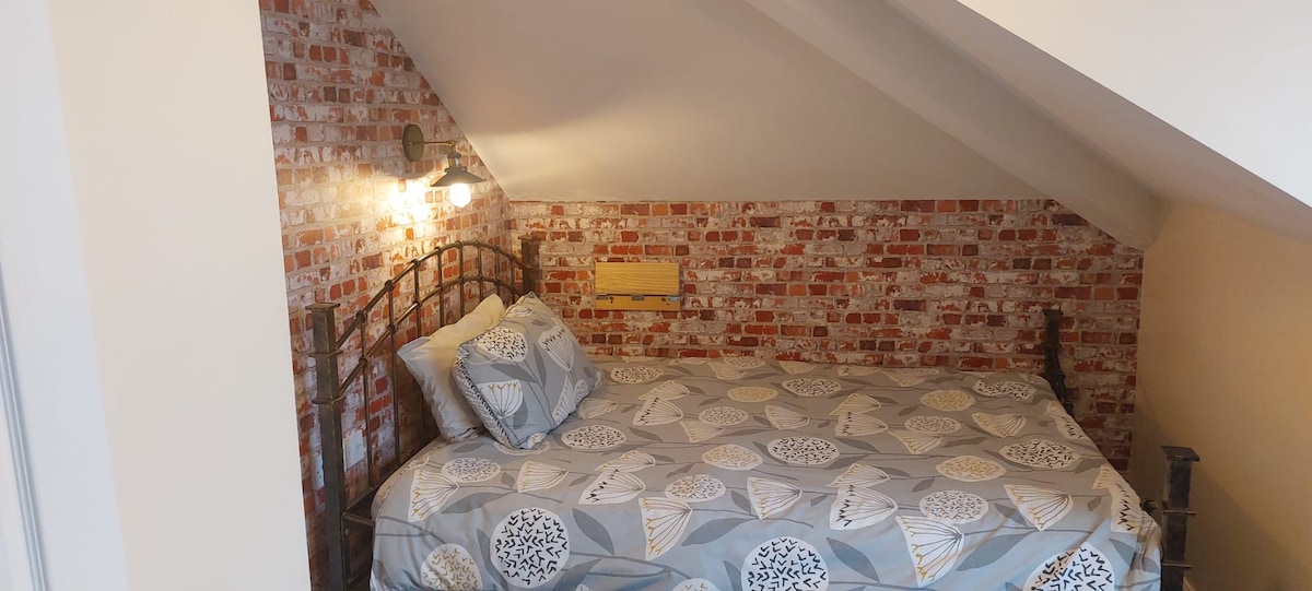 A cozy sleeping area is featured with a wrought iron bed, dressed in a patterned duvet cover. The exposed brick wall adds character to the space, with warm lighting provided by two bedside sconces, creating a comfortable atmosphere.