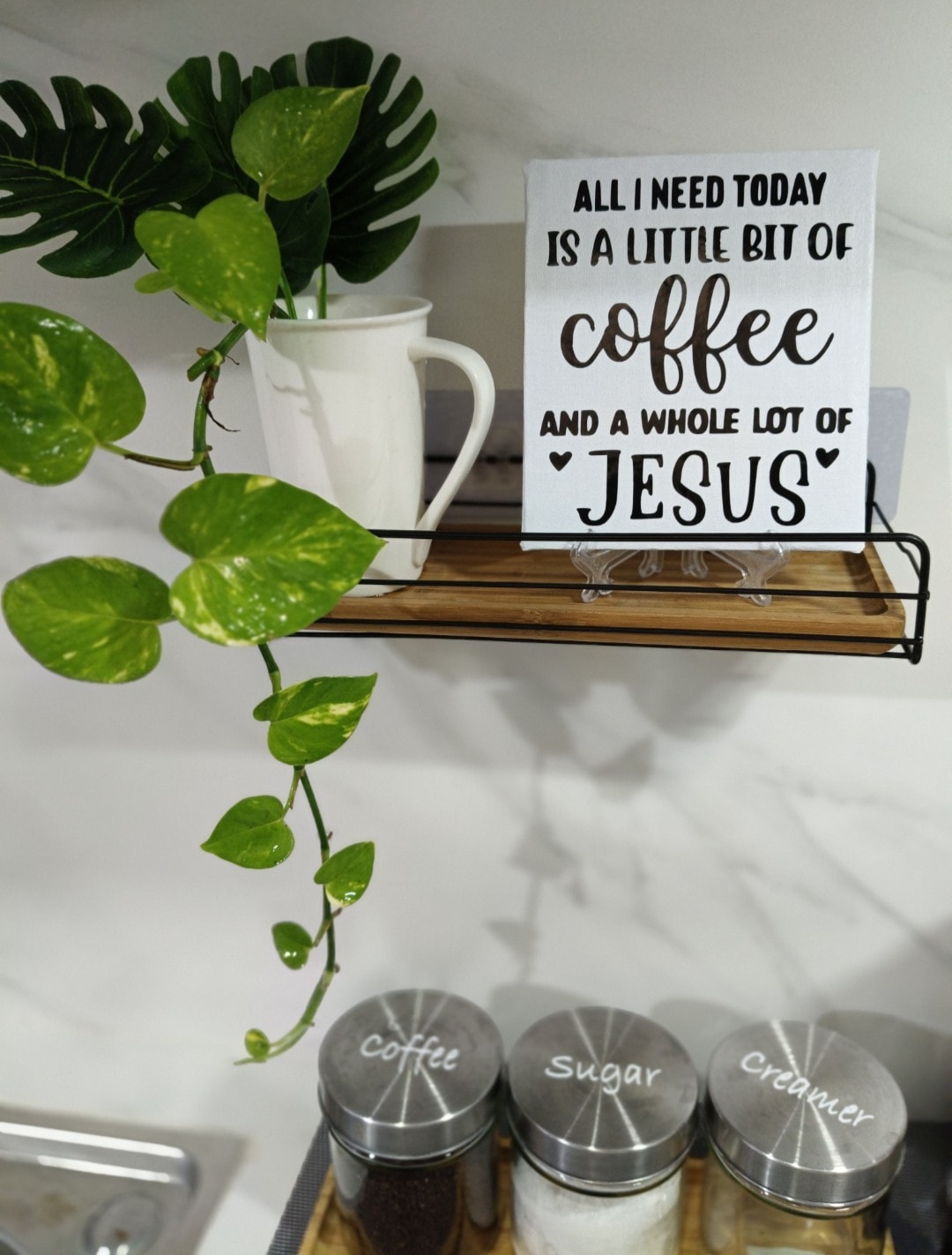 A shelf is displayed with a small white pitcher and a decorative sign featuring text about coffee and faith. Below, jars labeled 'Coffee,' 'Sugar,' and 'Creamer' are neatly arranged. A trailing green plant adds a touch of nature to the setting.