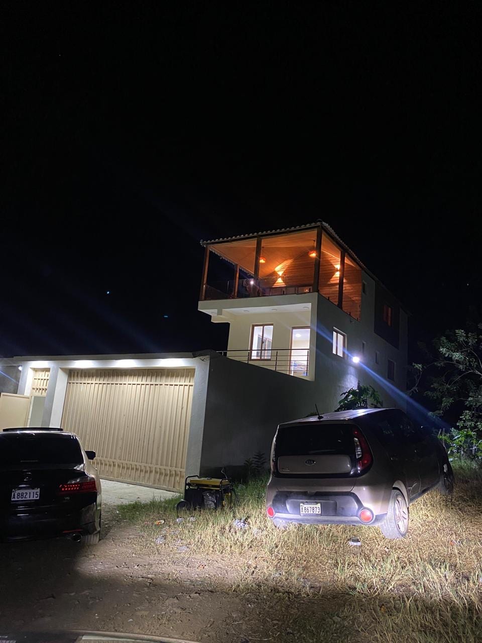 An illuminated two-story house stands at night, showcasing its distinct architecture. The upper level features a balcony surrounded by glass railing, while the lower level displays a garage with two parked vehicles. Ambient lighting highlights the house's contemporary design and natural surroundings.