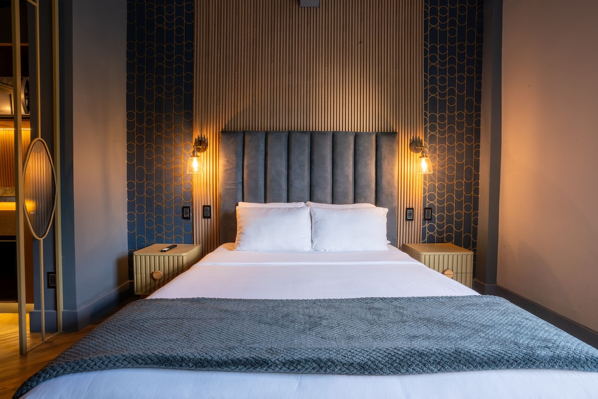 A modern sleeping space features a spacious bed framed by a textured headboard. Soft lighting is provided by wall-mounted fixtures on either side. Nightstands with a clean design complement the decor, while subtle patterns adorn the walls, creating a relaxed yet stylish ambiance.