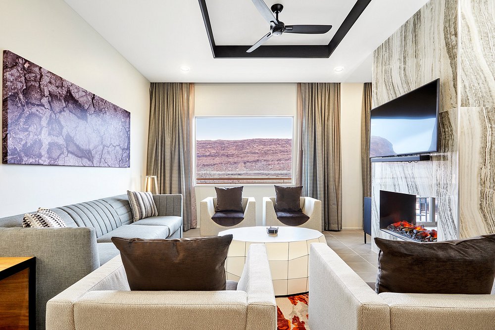 A modern living area is presented, featuring a sleek sofa and two stylish accent chairs. A round coffee table sits centrally, while a large window provides views of the red rock landscape. A fireplace and wall-mounted TV add comfort and convenience to the space.