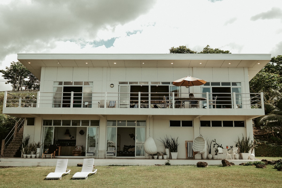 A modern two-story villa is presented, featuring large glass windows that offer views of the surrounding landscape. A spacious terrace is visible, shaded by an umbrella, while a well-maintained lawn with two lounge chairs can be seen in the foreground.