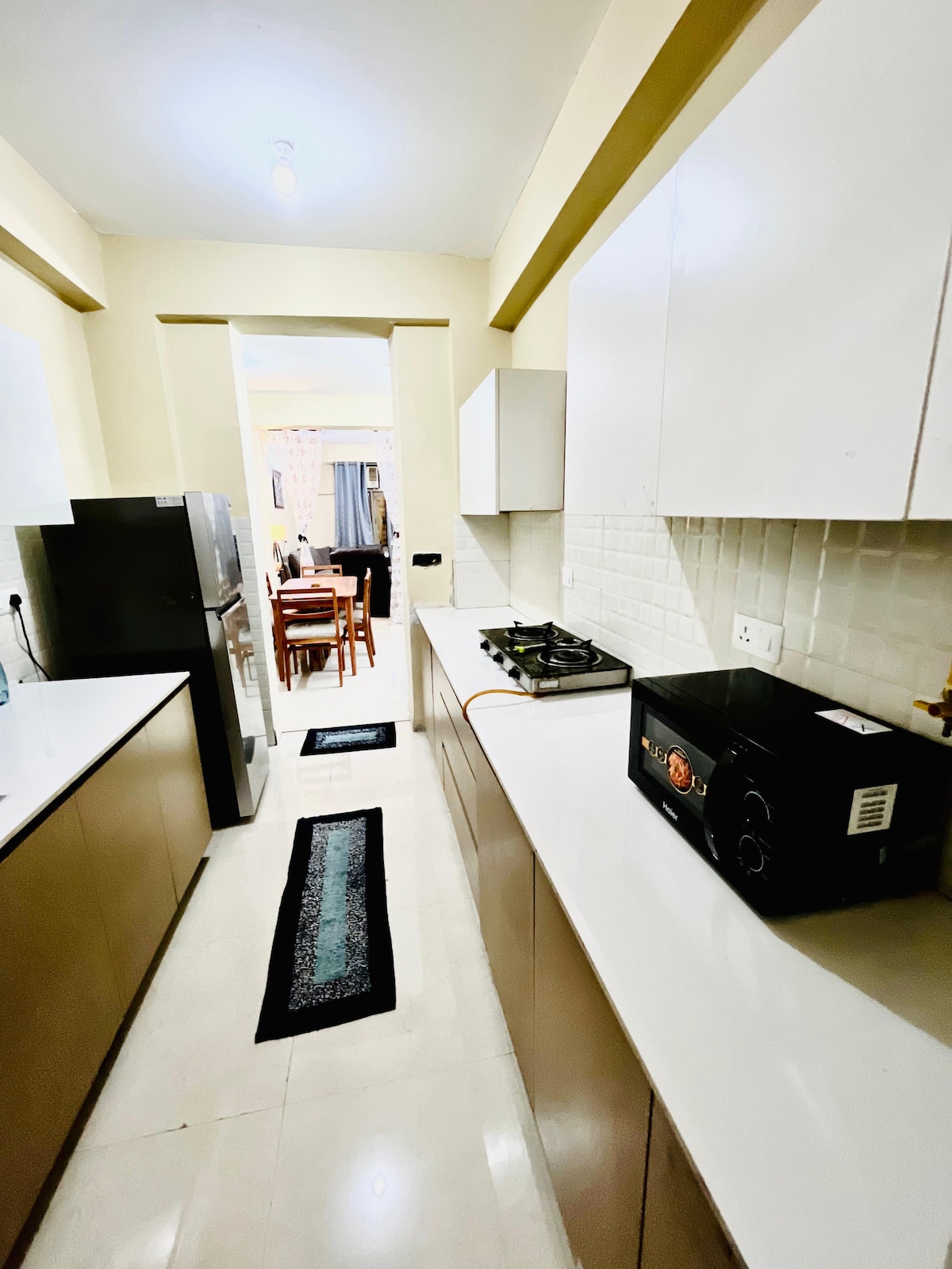 A well-organized kitchen features sleek cabinetry and modern appliances. A gas stove is positioned on the counter alongside a microwave. The space is complemented by a refrigerator, and a pathway leads to a dining area visible in the background.