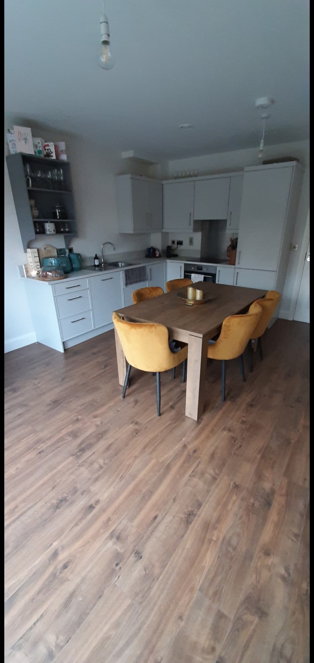 A modern kitchen features light grey cabinetry and a wooden dining table surrounded by four yellow chairs. A collection of kitchen essentials is displayed on a shelf, with additional counter space visible. Natural wood flooring adds warmth to the space.